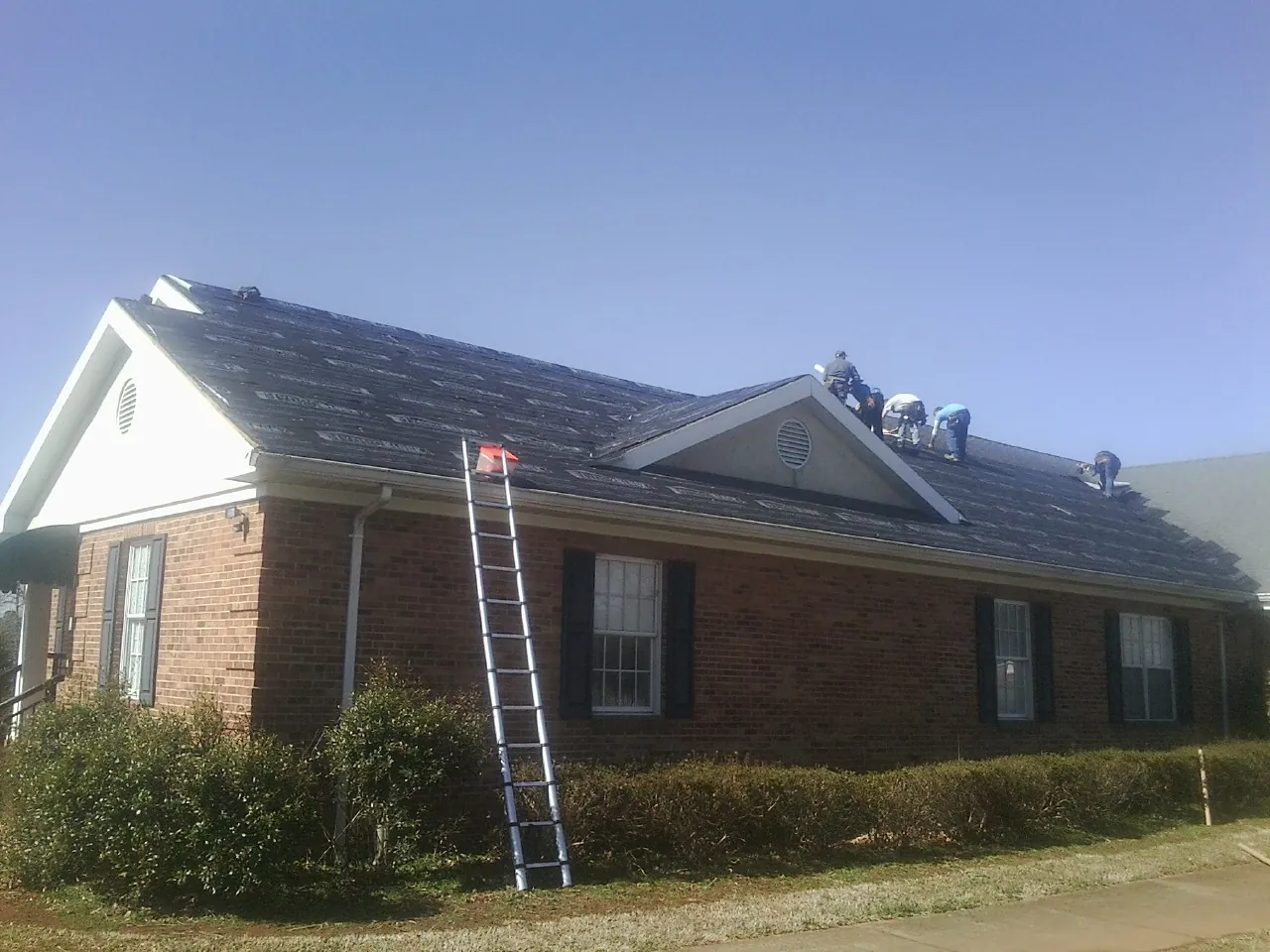 Crew tearing shingles off a brick ranch roof with a ladder set — Metal Roof Repair work by our crew in Gilbertville