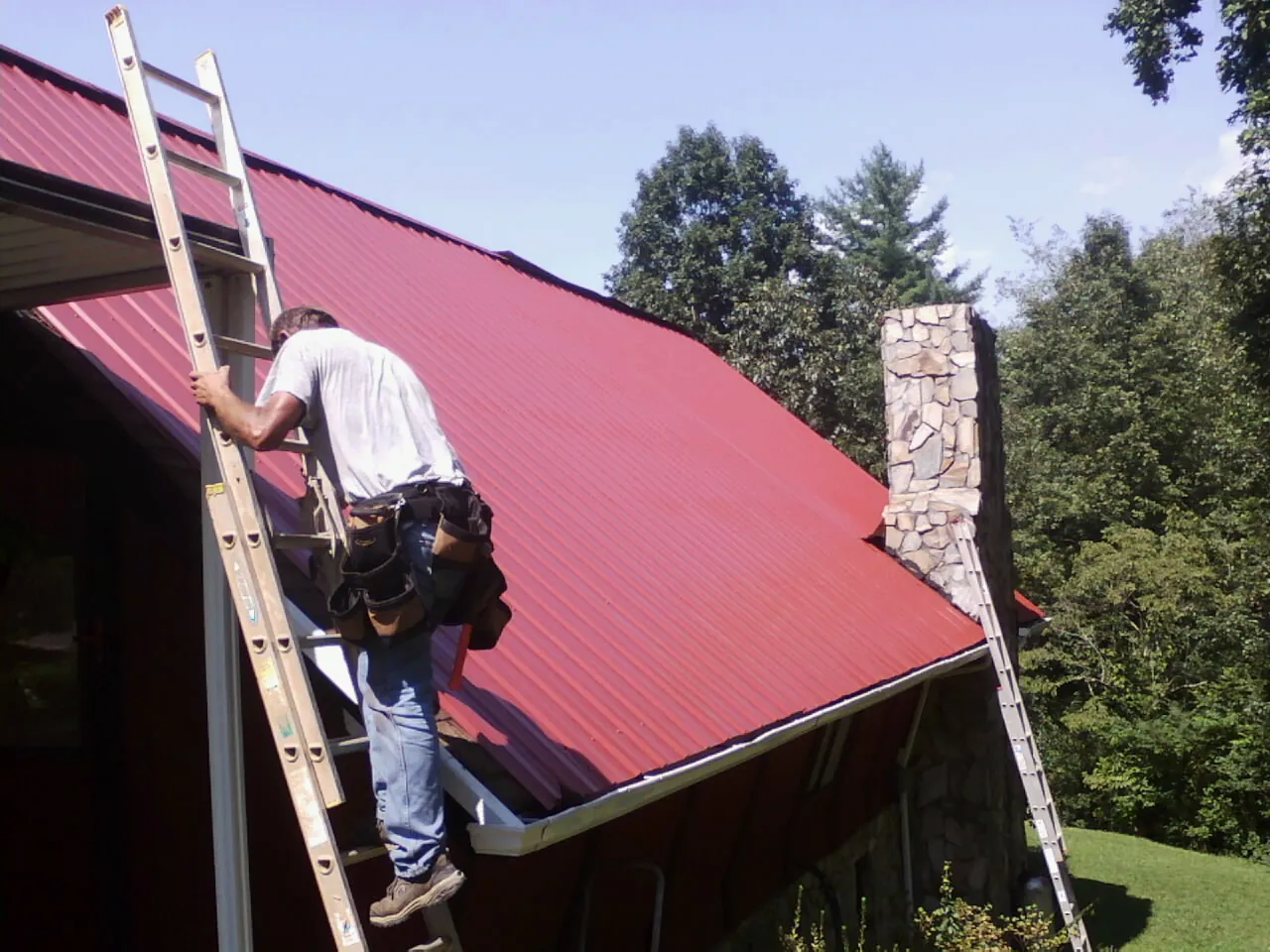 Roofer on a ladder beside a red metal roof and stone chimney — Soffit & Fascia Repair by Roofing Brothers in Gilbertville